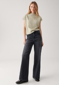 Femme se tenant debout dans un pantalon large noir, un haut sans manches clair et texturé, et des chaussures noires sur un fond clair uni.