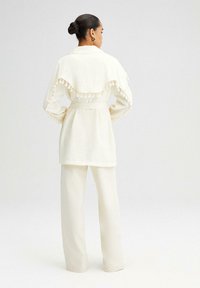 Conjunto color crema que presenta una chaqueta texturizada con volantes y acabados con borlas, además de pantalones de pierna ancha, mostrando un ajuste relajado.