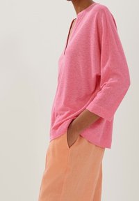 Rosa, texturerad långärmad skjorta med djup V-hals och sänkta axlar, ihop med ljusorange, löst sittande byxor.