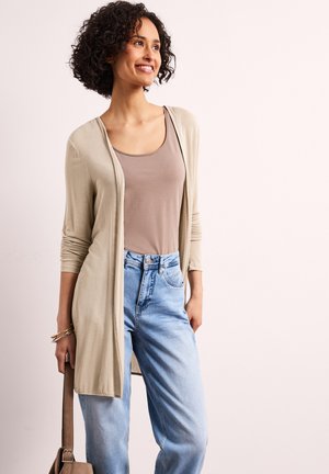 Kvinde smiler, iført beige åben cardigan, taupe tanktop, lyseblå jeans med høj talje, holder brun håndtaske ved siden.