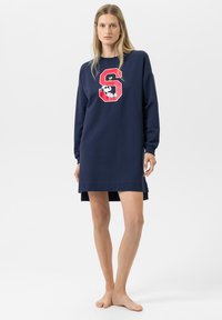 Vrouw staat blootsvoets in een marineblauwe jurk met lange mouwen, met een rode letter "S" en een Snoopy-afbeelding op de voorkant.