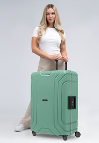 Valigia rigida di colore verde menta, con superficie testurizzata, multiple scanalature, manico retrattile, lucchetto a combinazione e quattro ruote per la mobilità.