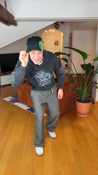 Must must müts rohelise lehe detailiga, hall kapuutsiga dressipluus varrukatel trükiga, hallid püksid ja valged tossud. Puidust põrand ja toataimed on nähtaval.