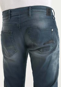 Jeans de mezclilla azul oscuro con un diseño desgastado, que cuentan con dos bolsillos traseros y un parche de cuero en la cintura como detalle de marca.