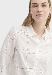 Chemise blanche présentant des motifs découpés complexes et un col, avec une patte de boutonnage et une texture de tissu douce.