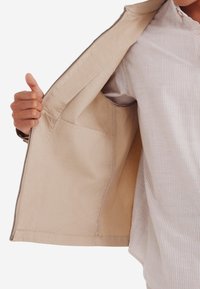 Beige jas met een rits aan de voorkant, met een gladde textuur en een binnenzak aan de linkerkant, gedragen over een gestreept shirt.