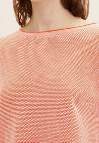 Personne portant un haut en tricot texturé orange et blanc avec un col rond et une coupe ample, visible du cou jusqu'au milieu du torse.