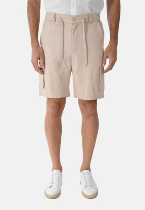 Uomo che indossa pantaloncini cargo beige con tasche laterali, una camicia bianca e sneaker bianche, in piedi davanti a uno sfondo bianco.