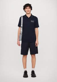 Mornarsko modra polo majica s kratkimi rokavi in kontrastnim ovratnikom; v kombinaciji z mornarsko modrimi kratkimi hlačami. Model drži svetlo sivo torbo z nastavljivim trakom.