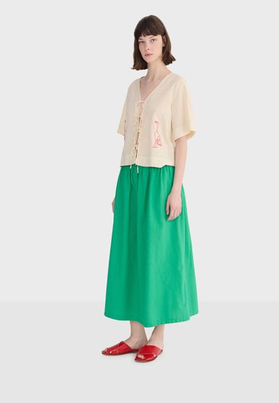 Femme portant un chemisier crème à manches courtes avec des nœuds à l'avant et des imprimés d'oiseaux rouges, une longue jupe verte et des sandales rouges à enfiler, debout immobile.