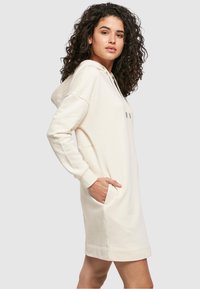 Hūdī tipa kleita gaiši krēmīgā krāsā, izgatavota no mīksta auduma. Aprīkota ar sānu kabatām, ribotām aprocēm un brīvu piegriezuma.