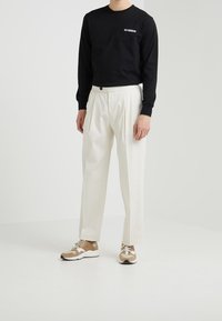 Svart långärmad skjorta med vit logotyp, krämfärgade veckade byxor och beige sneakers med vita detaljer. Minimalistisk design, avslappnad passform.