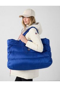 Sac fourre-tout bleu rembourré avec deux épaules épaisses, arborant un patch logo. Porté par une personne en veste blanche, mettant en valeur une texture douce et matelassée.