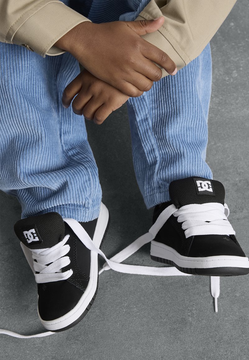 Zwarte DC sneakers met witte veters en logo, gedragen met lichtblauwe corduroy-broeken, rustend op een grijze ondergrond.