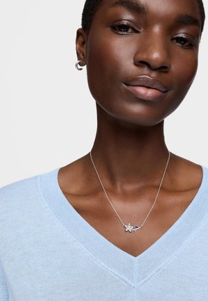 Mujer de piel oscura que lleva un suéter azul claro con cuello en V, un pendiente de aro plateado y un collar de plata con un colgante de flor y hoja púrpura.