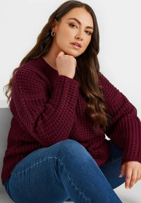 Burgundowy sweter dzianinowy o textured wzorze i szerokich rękawach, w połączeniu z klasycznymi niebieskimi dżinsami, siedzący na tle w jednolitym kolorze.