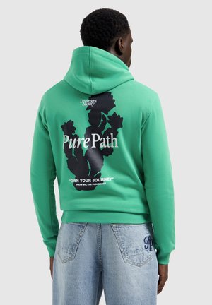 Person trägt grünen Hoodie mit schwarzem Kaktus-Design und Text "Pure Path", "Dreamers Society" und "Own Your Journey" auf der Rückseite, kombiniert mit hellen Jeans.