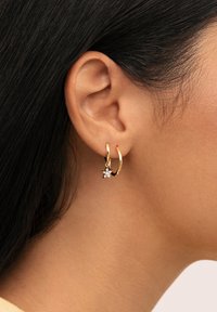 Pendiente de aro dorado con un pequeño charm en forma de estrella que presenta piedras transparentes, mostrado en una oreja, destacando su acabado metálico brillante y textura.