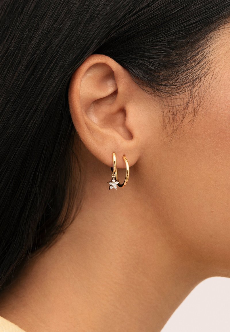 Pendiente de aro dorado con un pequeño charm en forma de estrella que presenta piedras transparentes, mostrado en una oreja, destacando su acabado metálico brillante y textura.