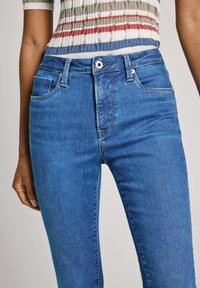 Los jeans de mezclilla azul presentan una silueta ajustada, un diseño de cinco bolsillos y un cierre frontal con botón y un acento metálico. Textura sutil de manchas.