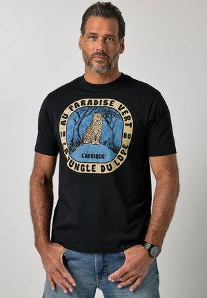 Mann mit Bart, der ein schwarzes T-Shirt mit Leopardengrafik und dem Text „Au Paradise Vert La Jungle Du Lopé L'Afrique“ trägt, steht mit den Händen in der Nähe der Taschen.
