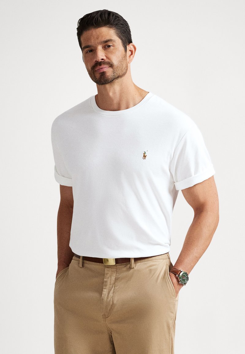 Vit t-shirt i bomull med uppkavlade ärmar och en liten logotyp på bröstet, kombinerad med beige byxor och ett brunt bälte. Enkel design.