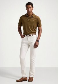 Καφέ πόλο μπλούζα, λευκά τζιν, καφέ ζώνη και καφέ παπούτσια. Φοράει ρολόι. Ο άνδρας στέκεται σε ουδέτερο φόντο, με τα χέρια στη μέση.