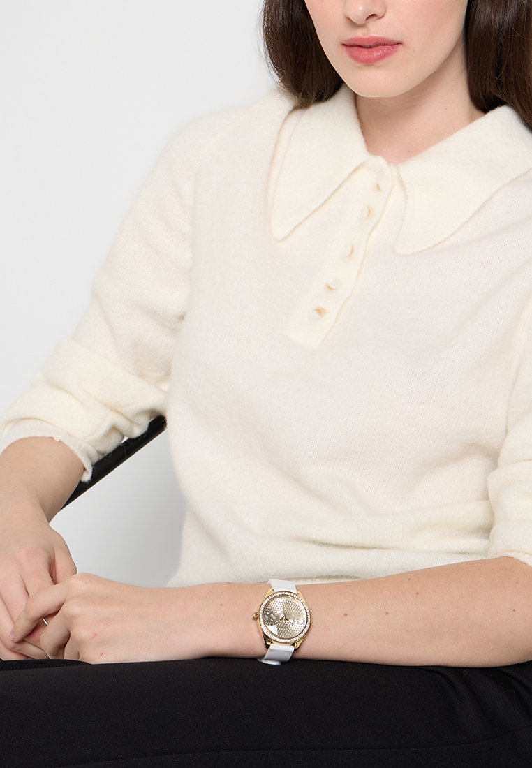 Suéter tejido de color crema con un gran cuello y carterilla con botones. Reloj de plata y oro en la muñeca, con esfera texturizada y correa blanca.