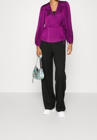 Blusa avvolgente in satin viola con maniche a puff lunghe, vita arricciata e pantaloni neri a gamba larga. Borsa metallica teal con tracolla a catena. Scarpe bianche.