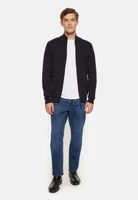 Svart zip-up jacka, vit t-shirt, blå jeans och svarta stövlar. Jackan har en hög krage och ribbade muddventiler.