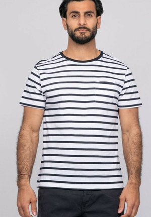 Homme aux cheveux courts et foncés et à la barbe, portant un t-shirt blanc à rayures horizontales noires avec une poche sur la poitrine, debout devant un fond clair.