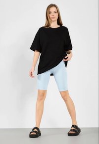 Mulher de pé contra um fundo branco, vestindo uma t-shirt preta oversized, calções de ciclismo azul claros e sandálias de plataforma pretas.