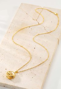 Gold pendant necklace with a round charm featuring floral design, hanging from an elongated, thin gold chain on a textured stone surface.