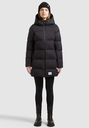 Femme portant un manteau matelassé noir à capuche, des leggings noirs, des bottes noires et un bonnet en laine noir, debout devant un fond uni.
