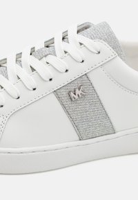 Baltos sportbačiai su sidabriniu tekstilės šoniniu panele, apvaliais raišteliais ir guminiu padu. Dėlionėje yra metalinė logotipo emblema šone ir tekstūruota viršutinė dalis.