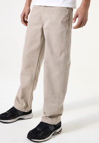 Beige Baumwollhosen mit geradem Schnitt, ausgestattet mit Fronttaschen und dezenten Knitterdetails, kombiniert mit schwarzen Sportschuhen.