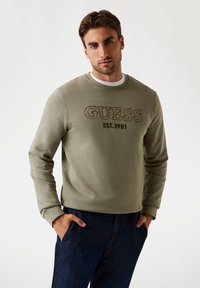 Felpa verde oliva in cotone, con collo a giro e polsini a costine, e il testo "GUESS EST.1981" ricamato sul davanti.