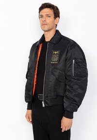 Giubbotto bomber in satin nero con fodera arancione, chiusura con zip, diverse tasche e una toppa in stile militare sul petto. Presenta polsini a coste.