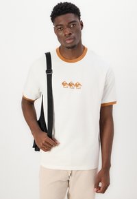 Camiseta blanca de manga corta con ribete marrón, que presenta un diseño gráfico. El modelo sostiene un bolso negro y lleva pantalones beige claros. Diseño simple y casual.