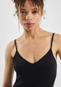 Vrouw met krullend haar, gekleed in lange, hangende oorbellen en een zwarte mouwloze top met dunne bandjes, die rechtop staat tegenover een effen achtergrond.