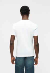 Άνδρας με κοντά σγουρά μαλλιά, που φοράει απλό λευκό κοντομάνικο μπλουζάκι και ξεθωριασμένο μπλε τζιν, στέκεται με την πλάτη προς το λευκό φόντο.