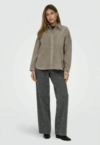 Corduroy shirt jacket in taupe with a button closure and two chest pockets, paired with black jeans featuring scattered white embellishments.