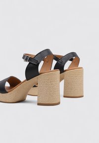 Sandalias de tacón de piel negra texturizada con un ancho tacón de bloque de paja trenzada y correa de tobillo ajustable. Diseño de punta abierta con una forma curva.