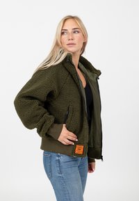 Oliwkowa kurtka sherpa z ribbowanym dołem i mankietami, bocznymi kieszeniami oraz pomarańczową metką po lewej stronie. Zamek błyskawiczny z przodu.