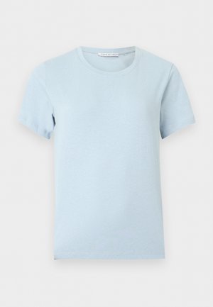 Noor naine pikkade tumedate juustega, kellel on seljas helesinine lühikeste varrukatega T-särk, helesinised teksad musta vööga ja mustad kontsaga sandaalid, seisab vaba poosiga.