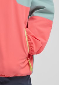 Leichte Jacke in Korallen- und Tealtönen mit einer Reißverschlusstasche und gelben Beschlägen. Glatte Textur und Color-Block-Design.