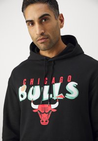 Felpa nera con cappuccio caratterizzata da una grafica rossa e teal. Il design include il testo "CHICAGO BULLS" e un'illustrazione della testa di un toro.