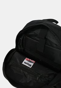 Zaino nero con un compartimento principale con cerniera, caratterizzato da un interno liscio, una tasca con cerniera e un'etichetta con accenti rossi e blu.