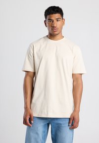 Beige puuvillane t-särk lühikeste varrukatega ja ümarate kaelustega, näidatud mudeli seljas lihtsa tausta ees.