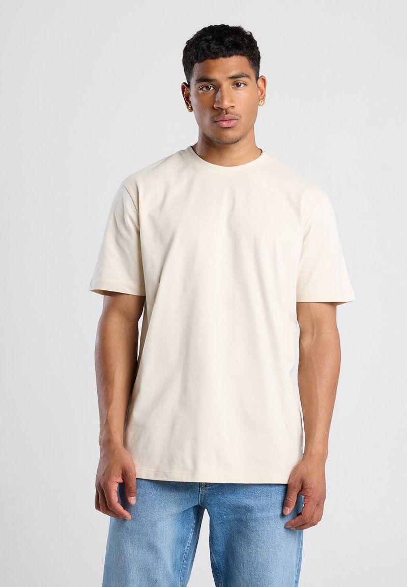 Beige puuvillane t-särk lühikeste varrukatega ja ümarate kaelustega, näidatud mudeli seljas lihtsa tausta ees.